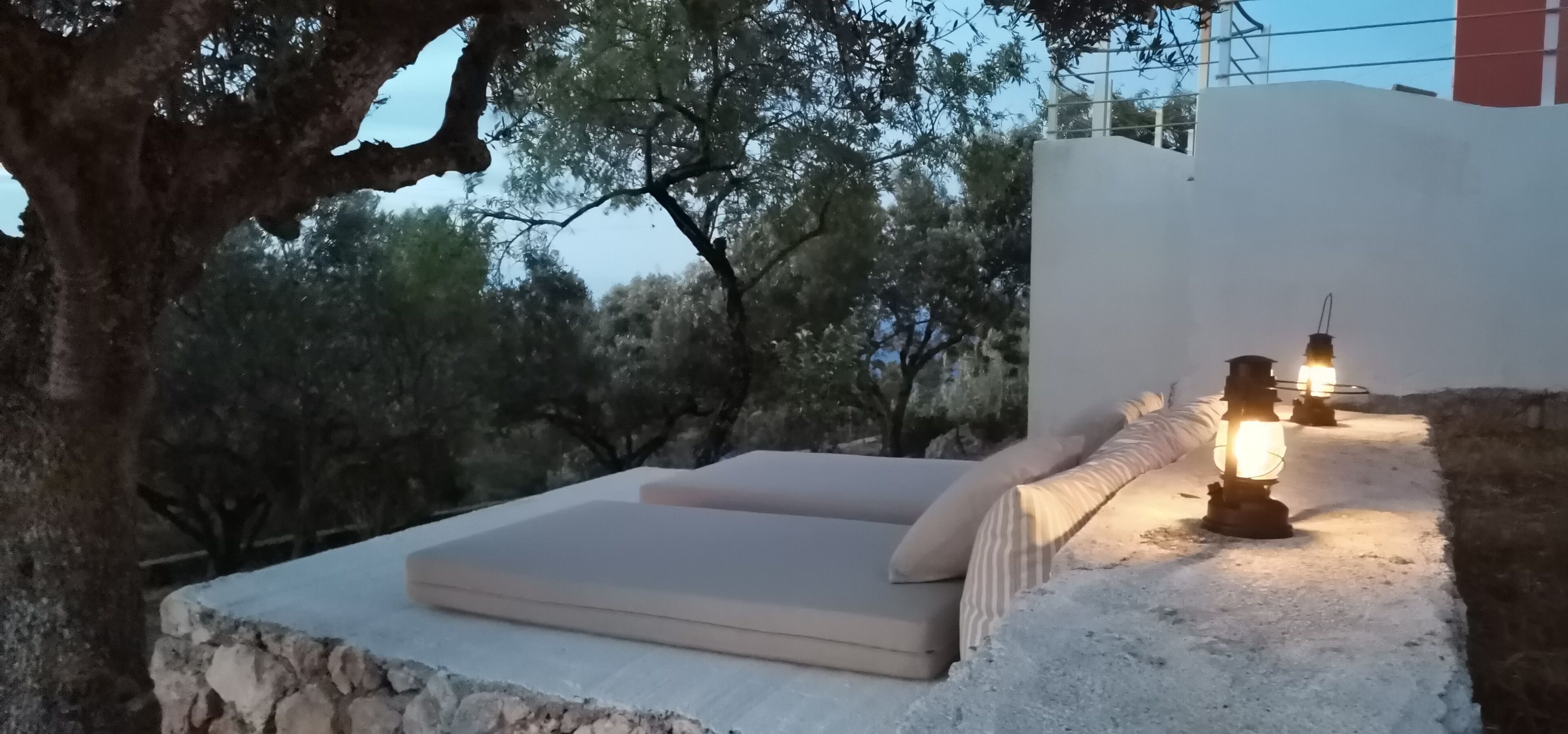 A stone bench with cushions, a lantern, and a view of trees and a building in the background.