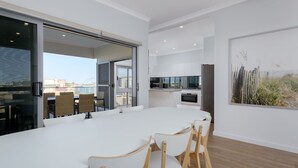 A modern dining area with a large table and chairs, a kitchen in the background, and a large window with a view of the city. - 22 Cooyou Close - Perfect Little Get Away (Exmouth)