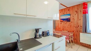 Una cucina con mobili bianchi, un lavello nero, una macchina da caffè e un tostapane. Si nota una parete in legno con un quadro e una tenda rossa.