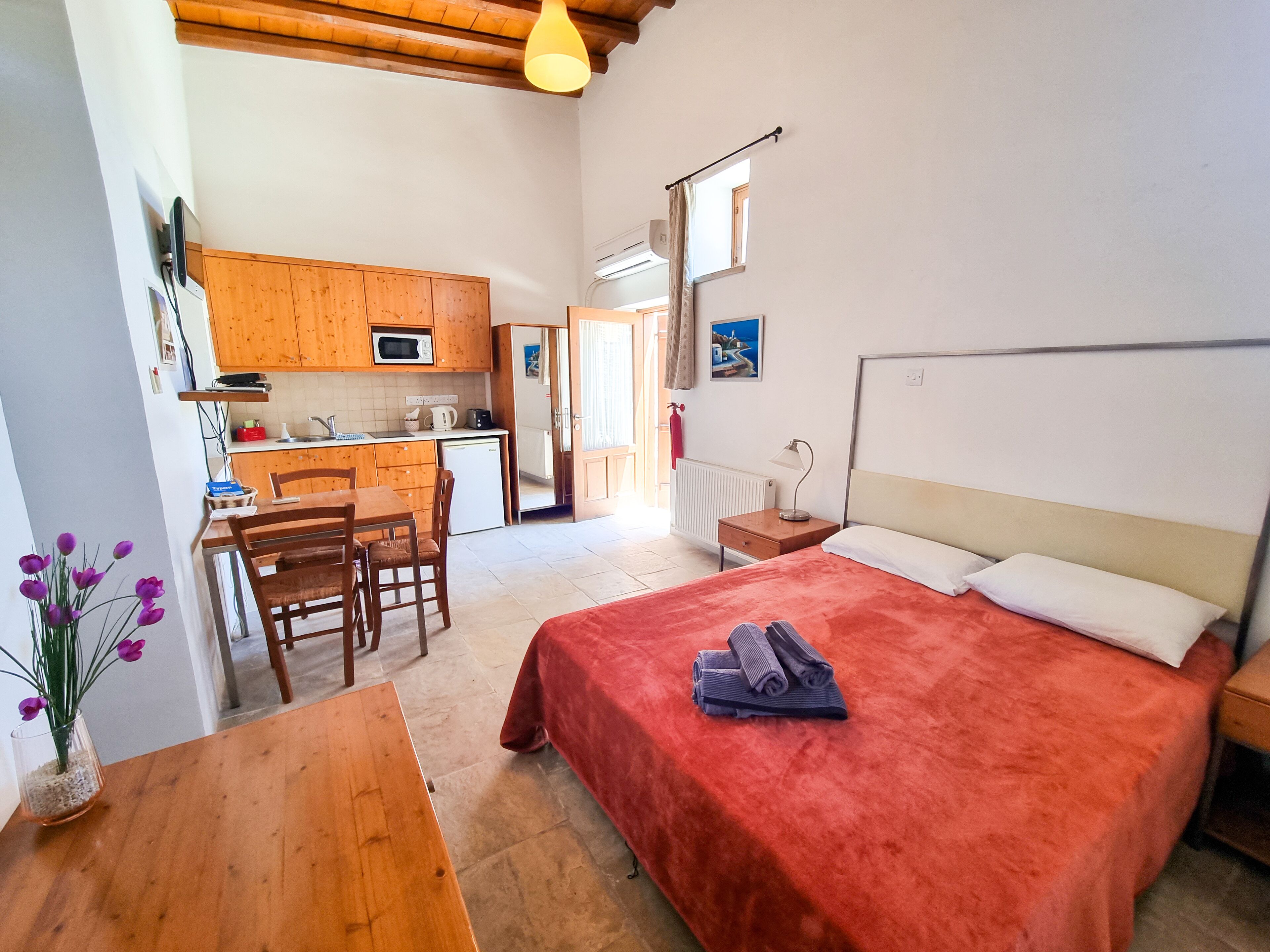 Een kleine kamer met een bed, een bureau, een stoel, een houten tafel en een kitchenette met een magnetron en een koelkast.