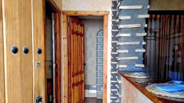 Uma porta de madeira com puxador de metal e fechadura, dando acesso a um corredor revestido de azulejos com parede de pedra e uma janela.