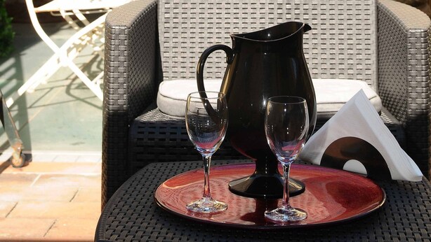 Un patio al aire libre con mobiliario de mimbre, una mesa dispuesta con una jarra y copas de vino, y una vista de un tejado de baldosas al fondo.