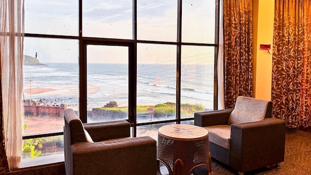 A room with a large window, two armchairs, a small round table, and patterned curtains. - SUKHSAGAR BEACH RESORT Kunkeshwar  (Devgad)