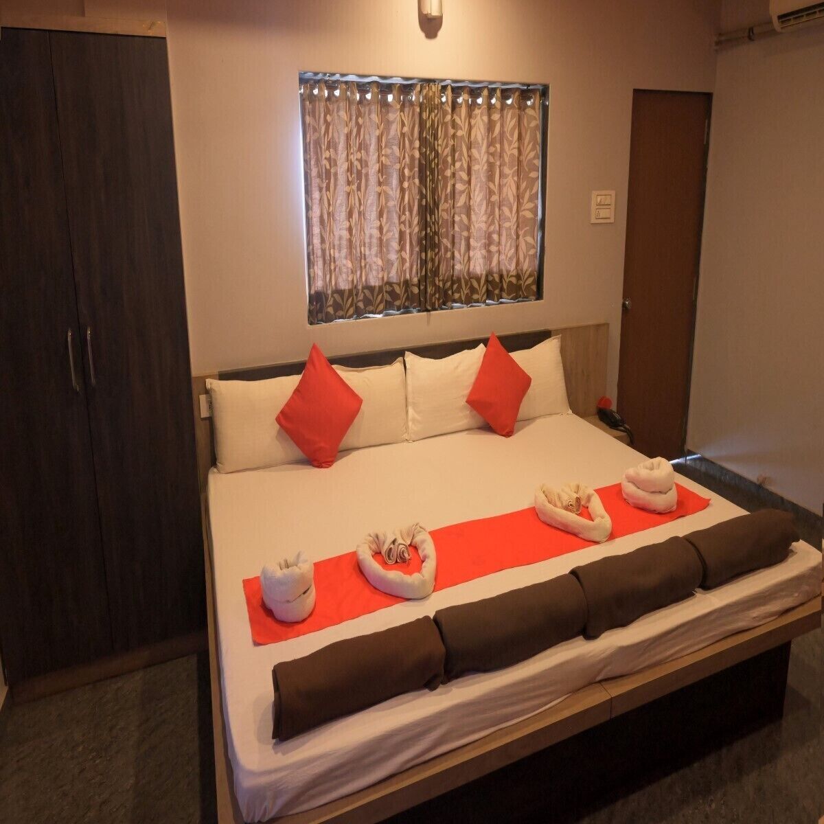 A neatly made bed with red and white pillows, a red blanket, and a set of towels on a red mat.