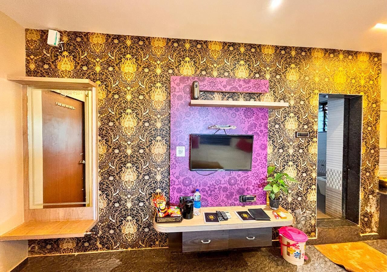 A hotel room with a patterned wallpaper, a television mounted on the wall, a small table with a plant, and a door.