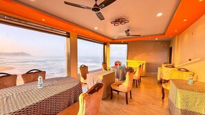 A dining area with tables and chairs, a ceiling fan, and large windows overlooking a landscape. - SUKHSAGAR BEACH RESORT Kunkeshwar  (Devgad)