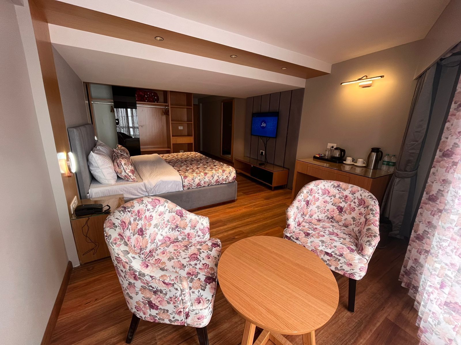 A compact hotel room with a bed, two floral armchairs, a small wooden table, a TV, and a kitchenette.