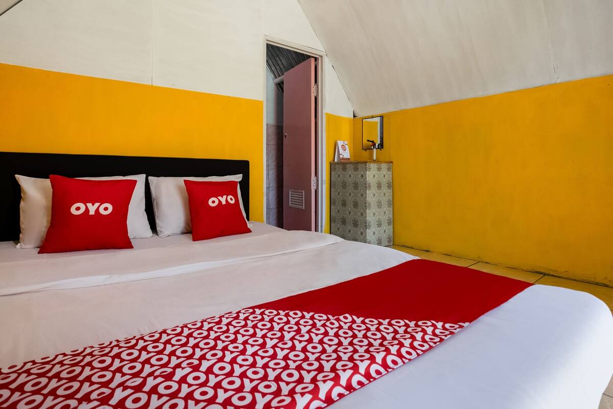 A hotel room with a bed, two red pillows, a red and white patterned blanket, a wardrobe, and a mirror on the wall.
