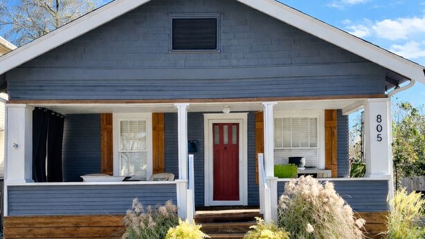 A single-story house with a red front door, a porch with white railings, and a small front yard with plants.