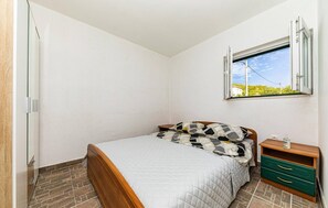 Quarto com cama, mesa de cabeceira, janela e prateleira fixada na parede.