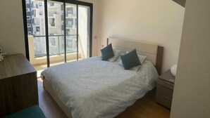 A bedroom with a large bed, a nightstand, a wooden desk, and a sliding glass door leading to a balcony.