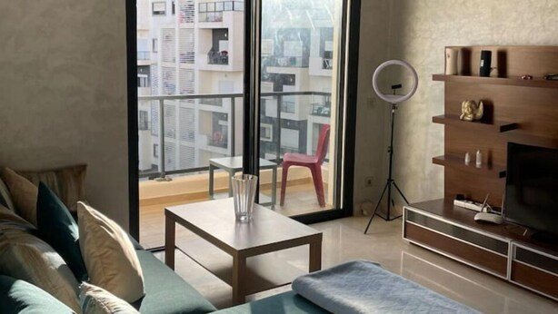A modern living room with a sofa, a small table, a TV stand, and a balcony with a view of other buildings.