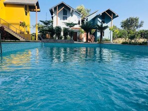 Una piscina con superficie de agua azul cristalina, rodeada de edificios modernos con fachada amarilla y blanca, y una escalera que conduce a un balcón.