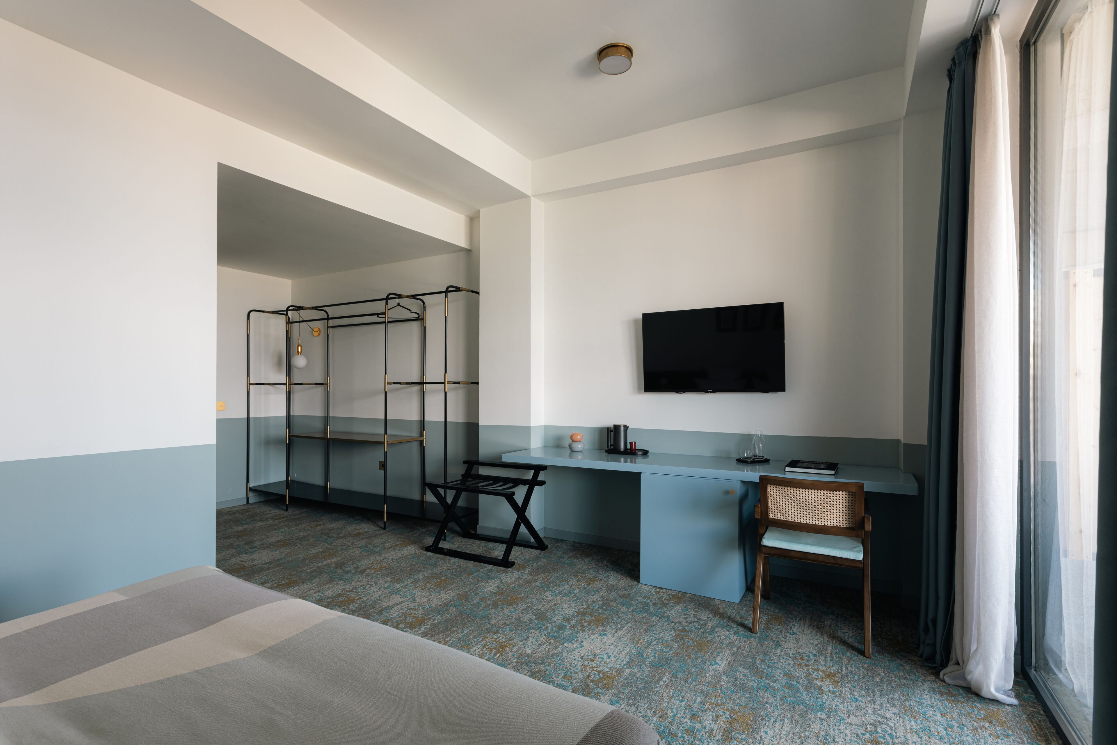 Une chambre d’hôtel comprenant un lit, un bureau avec une chaise, un téléviseur fixé au mur et une fenêtre avec des rideaux.