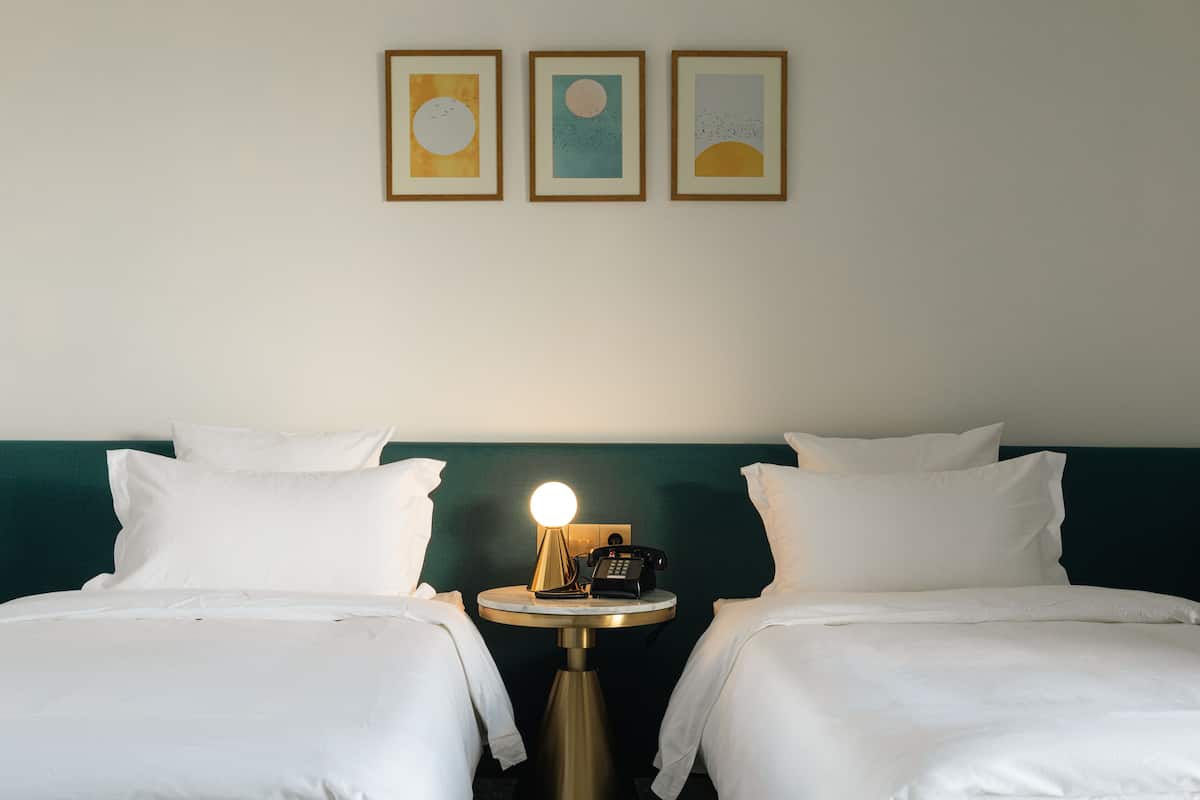 A hotel room with two beds, a bedside table with a lamp, a phone, and three framed artworks on the wall.