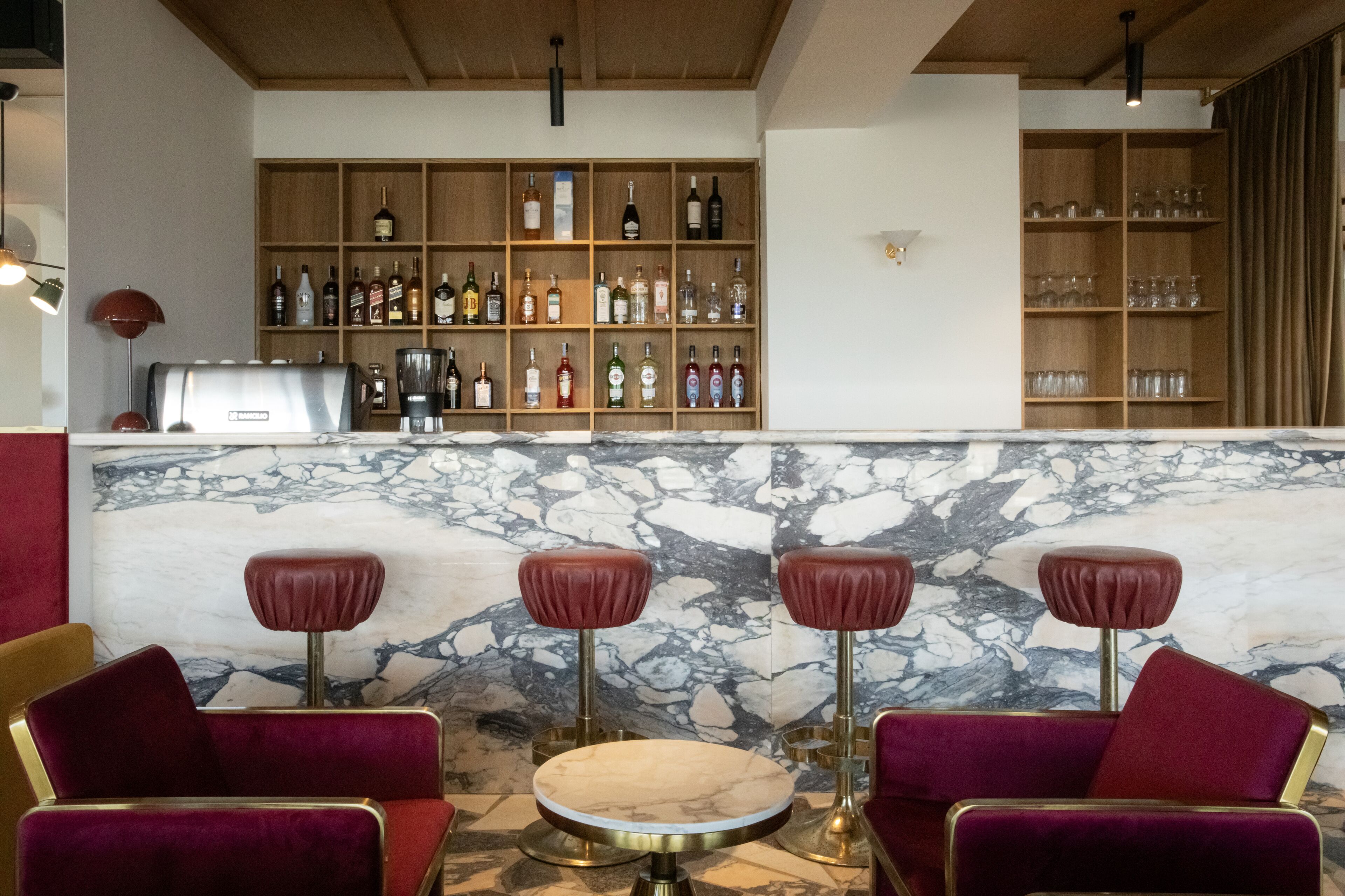 A bar area with marble countertops, red bar stools, and shelves stocked with bottles and glasses.