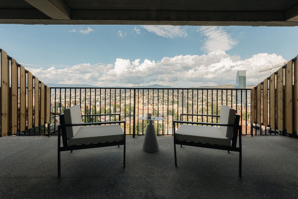 A balcony with two chairs and a small table, overlooking a cityscape with mountains and a tall building.