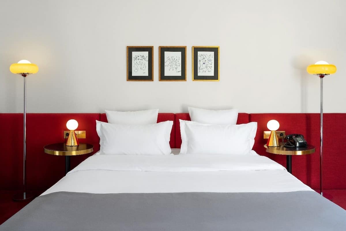 A hotel room with a bed, two bedside tables with lamps, a red accent wall, and three framed artworks on the wall.