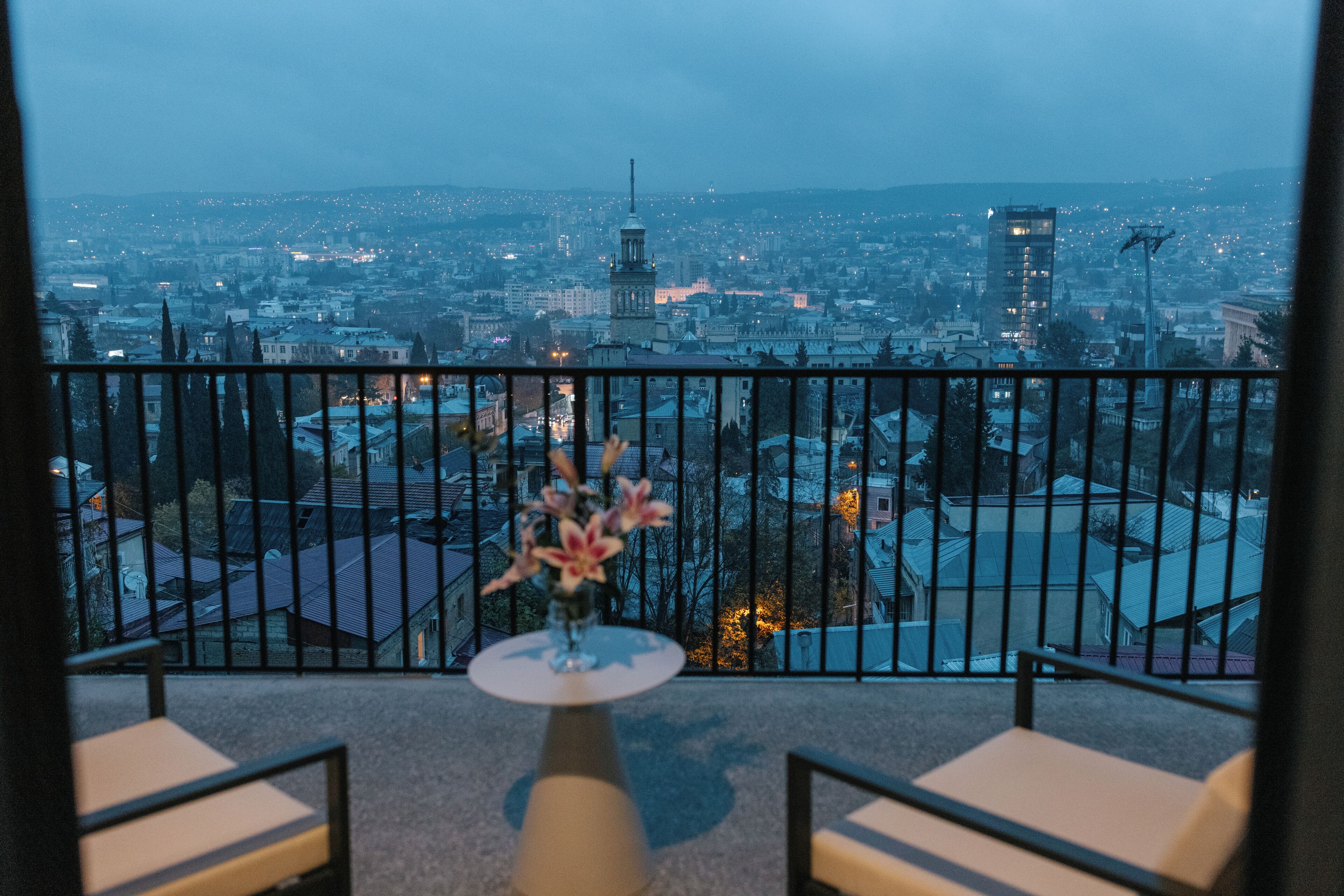 A balcony with a view of a cityscape, a table with a vase of flowers, and two chairs.