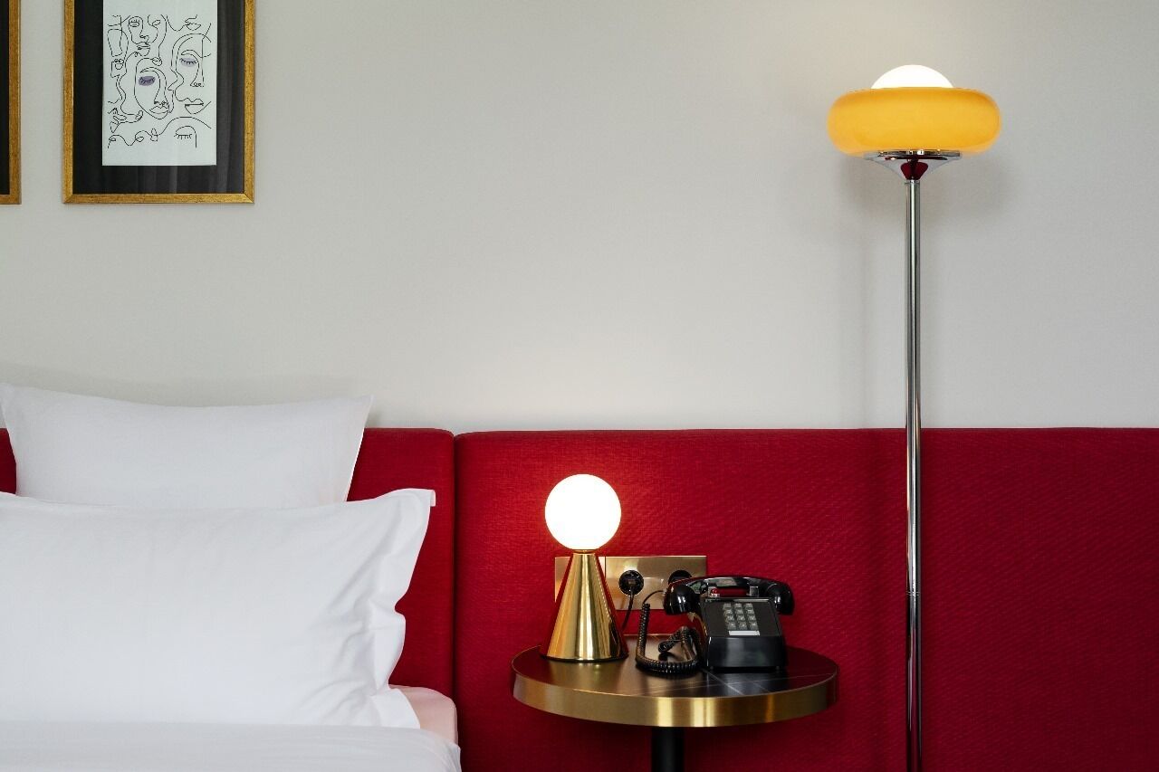 A hotel room with a red headboard, a bed with white bedding, a bedside table with a lamp, and a framed artwork on the wall.