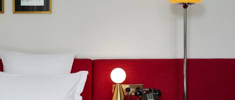 A hotel room with a red headboard, a bed with white bedding, a bedside table with a lamp, and a framed artwork on the wall.