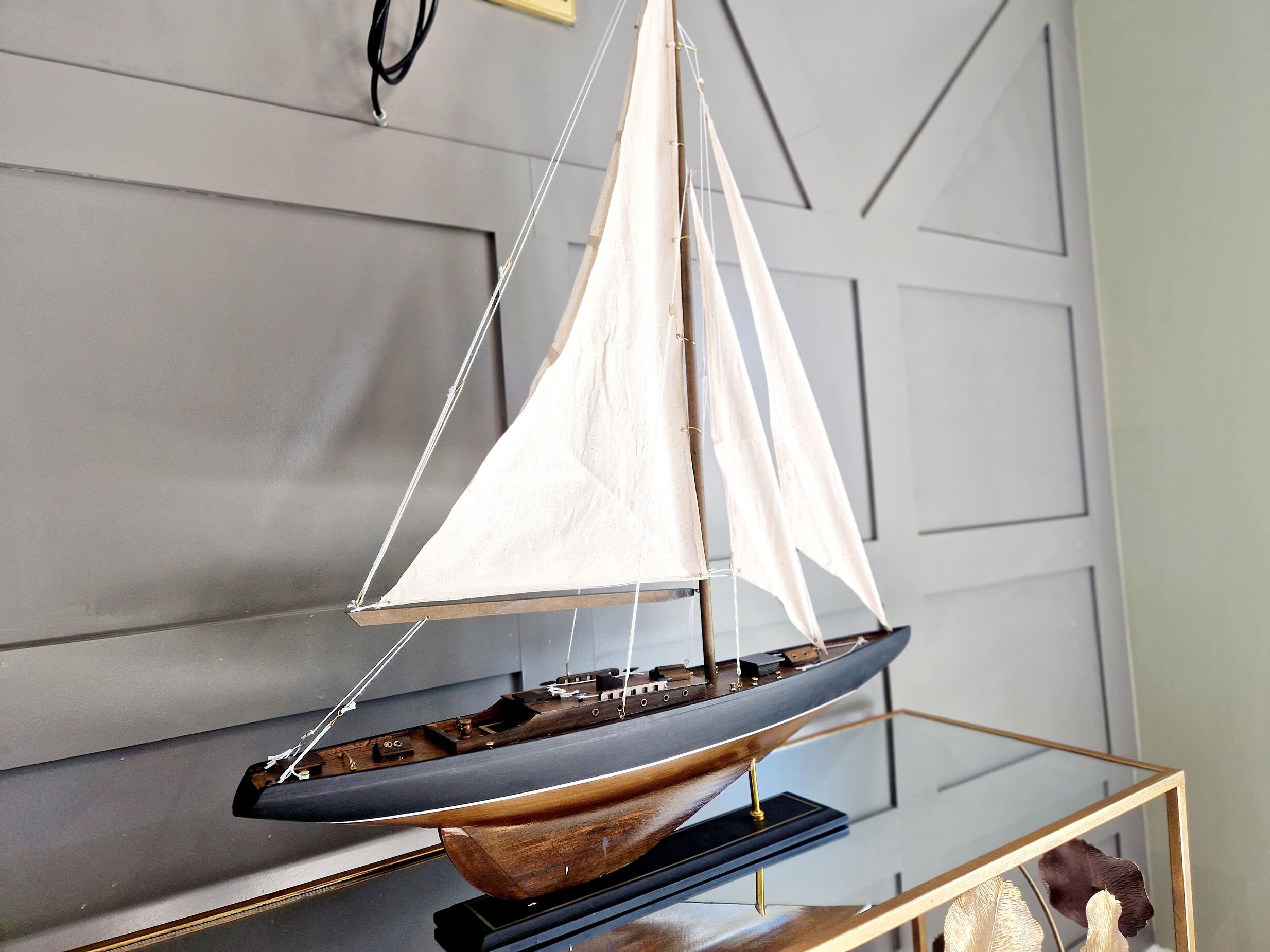 A model sailboat displayed on a glass table against a grey wall.