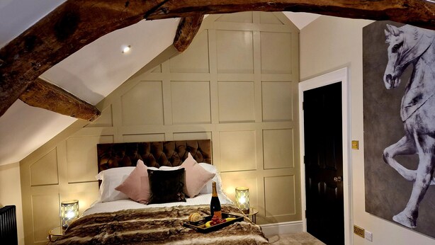 A bedroom with a large bed, a wooden ceiling beam, a headboard, a wall-mounted painting, and a nightstand with a bottle and fruit. - The Cruck (Preston)