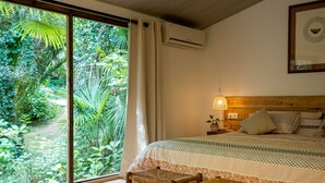 A bedroom with a large window, a bed with patterned bedding, a bedside lamp, and a small wooden stool.