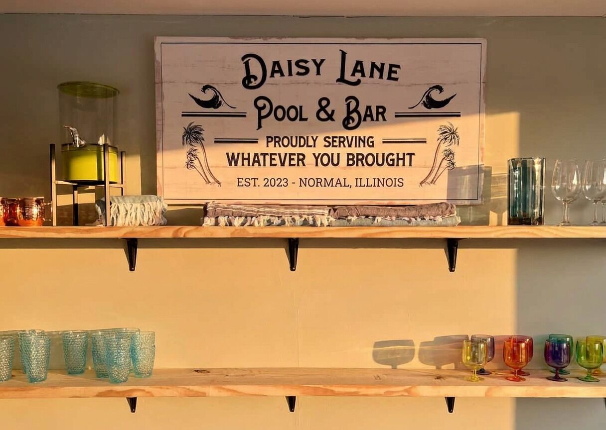 Un letrero para Daisy Lane Pool & Bar con un diseño tropical, un estante con vasos de colores y un recipiente de vidrio a la izquierda.