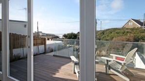 Une terrasse en bois avec une balustrade en verre, une chaise blanche et une vue sur une colline parsemée de maisons.