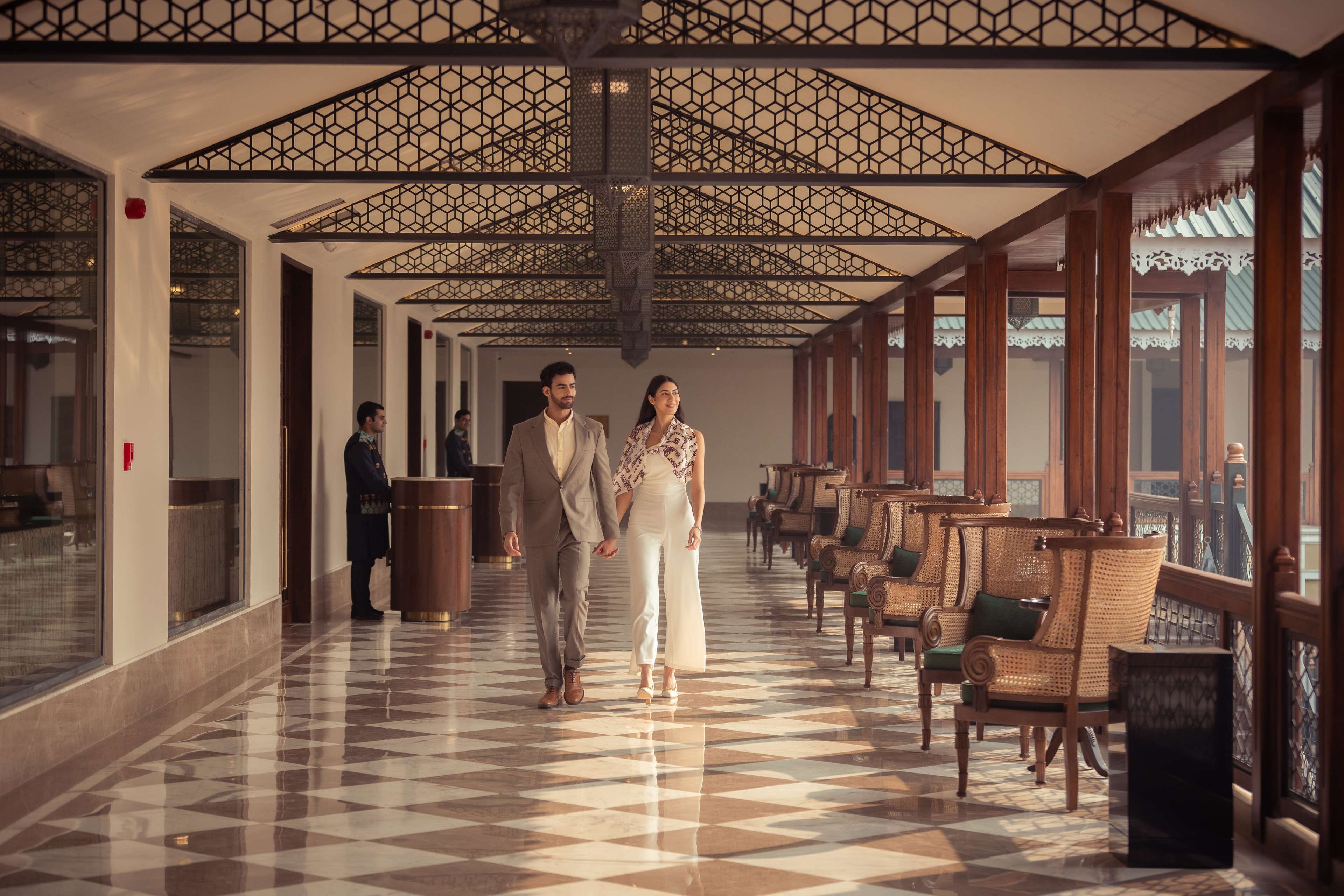 A hotel corridor with a checkered floor, wooden chairs, and a patterned ceiling.