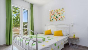Un dormitorio con cama, cortinas verdes, ventana con vistas, una mesita redonda con lámpara y una obra de arte colorida en la pared.