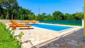 Una piscina con patio de piedra, dos sillas reclinables y un muro de piedra.