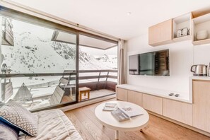 A modern hotel room with a large sliding glass door to a balcony, a flat-screen TV mounted on the wall, a small wooden table, and a sofa.