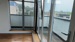 A room with a large sliding glass door leading to a balcony, wooden flooring, and a view of buildings in the distance. - Impeccable 2 Bed 2 Bath Penthouse (London)