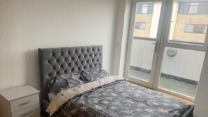 A bedroom with a tufted headboard, a patterned bedspread, a nightstand, and a view through a sliding glass door to the outside. - Impeccable 2 Bed 2 Bath Penthouse (London)