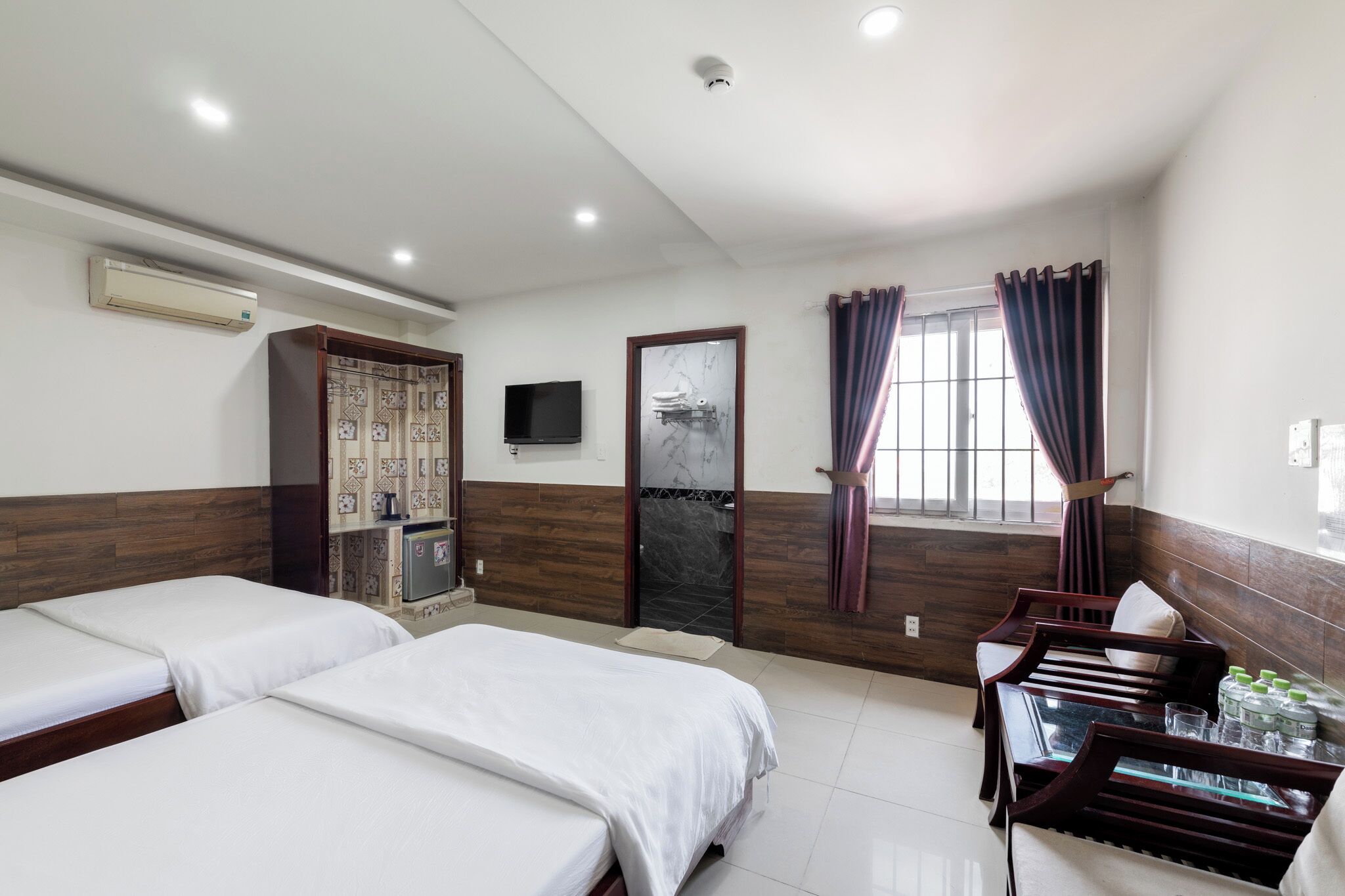 A hotel room with two beds, a wooden wardrobe, a television, and a dining area with chairs and a table.