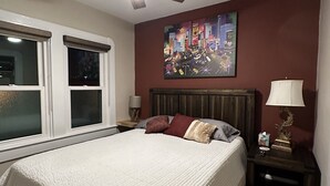 Un dormitorio con cama, ventilador de techo, mesita de noche, lámpara y una pintura de paisaje urbano en la pared.
