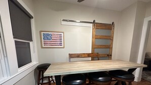 Un comedor con una mesa de madera y cuatro taburetes, una ventana con persianas, una bandera estadounidense enmarcada y una escalera de madera apoyada en la pared.
