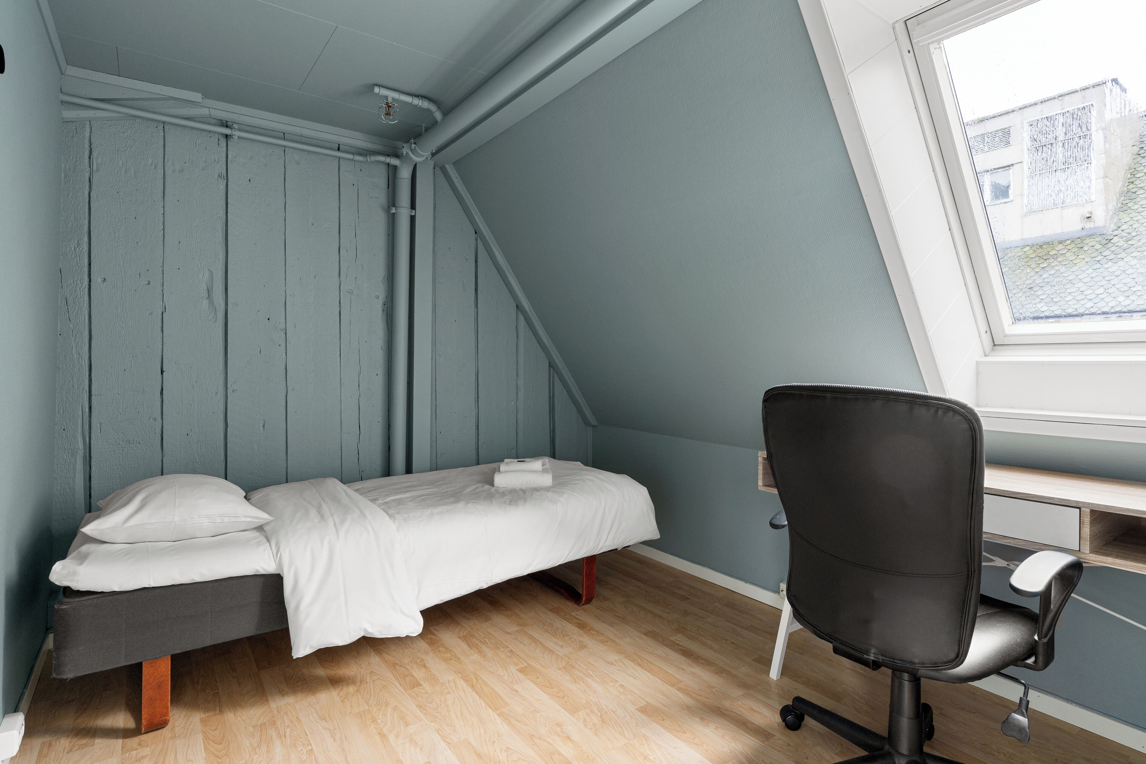 A small, single-bed room with a desk and chair, a sloped ceiling, and a window with a view of buildings.