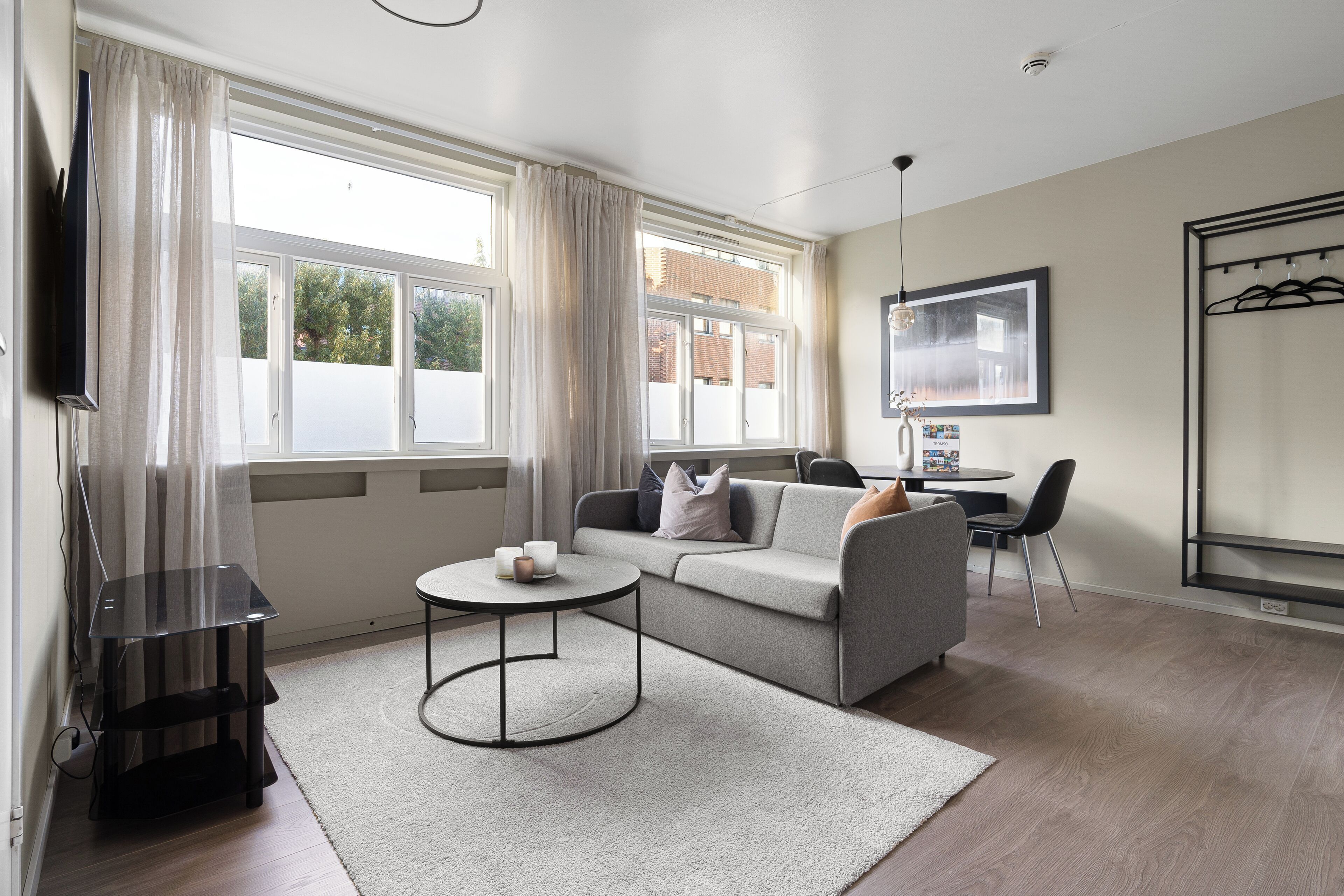 A modern living room with a sofa, a coffee table, and a TV. There is a window with curtains, a coat rack, and a framed picture on the wall.