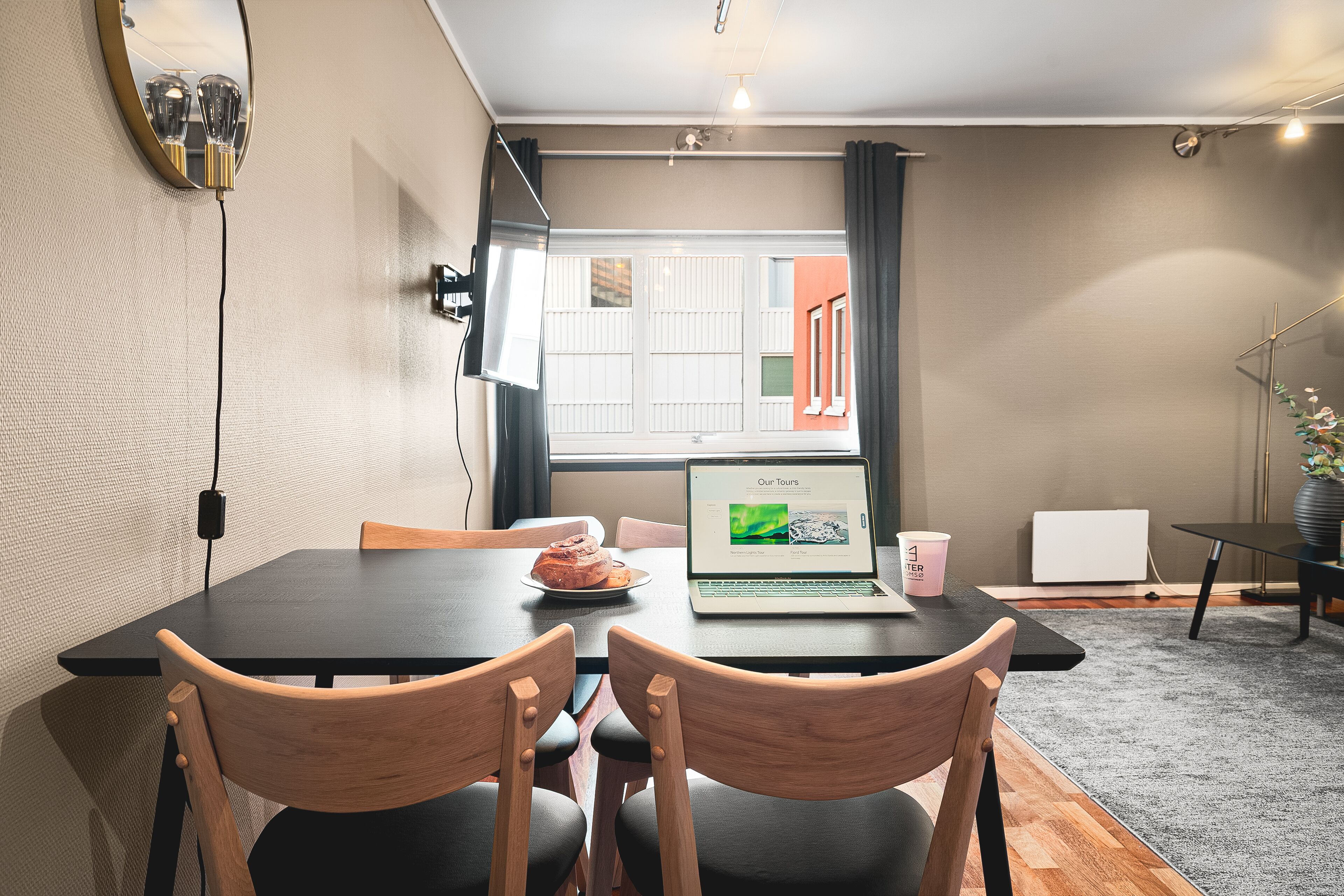 Un comedor moderno con una laptop sobre una mesa, dos sillas de madera, un plato de comida y una ventana con persianas.