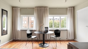 A room with a round table, two chairs, a window with curtains, and a view of buildings outside. - Amalie Apartments By First (Tromsø)