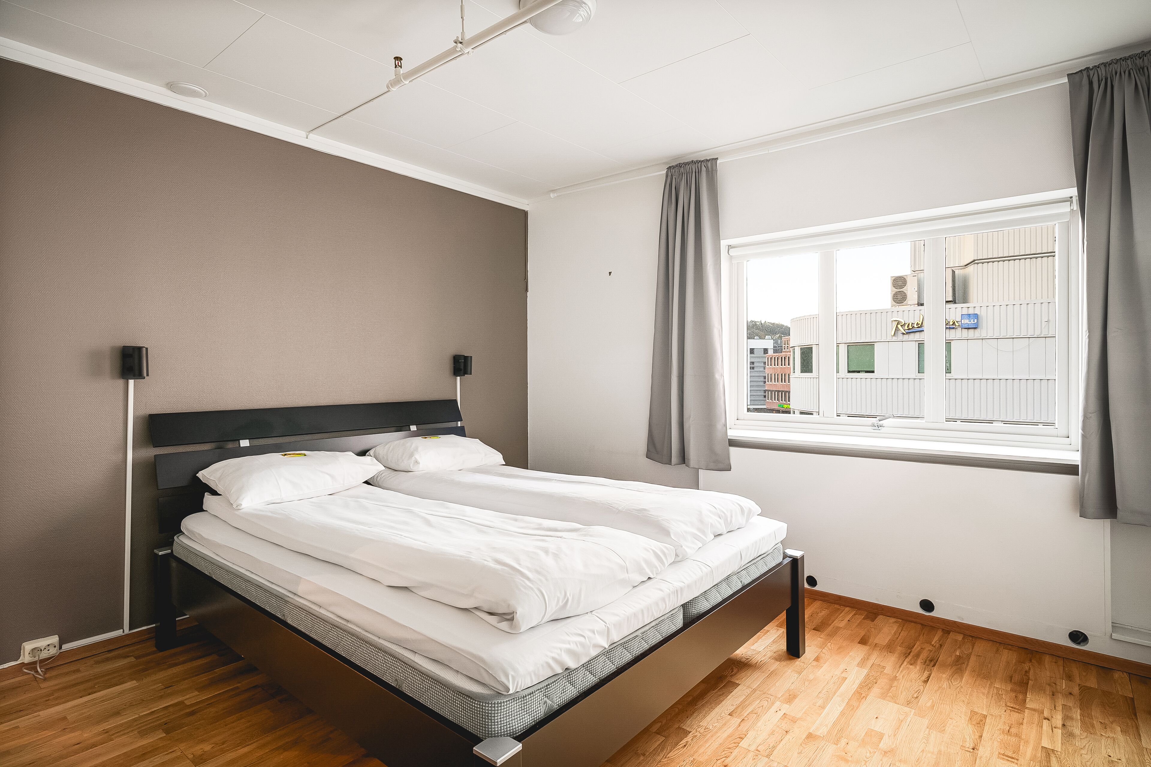 Ein modernes Schlafzimmer mit einem Bett, Nachttischen, einem Fenster mit Vorhängen und Blick auf Gebäude.