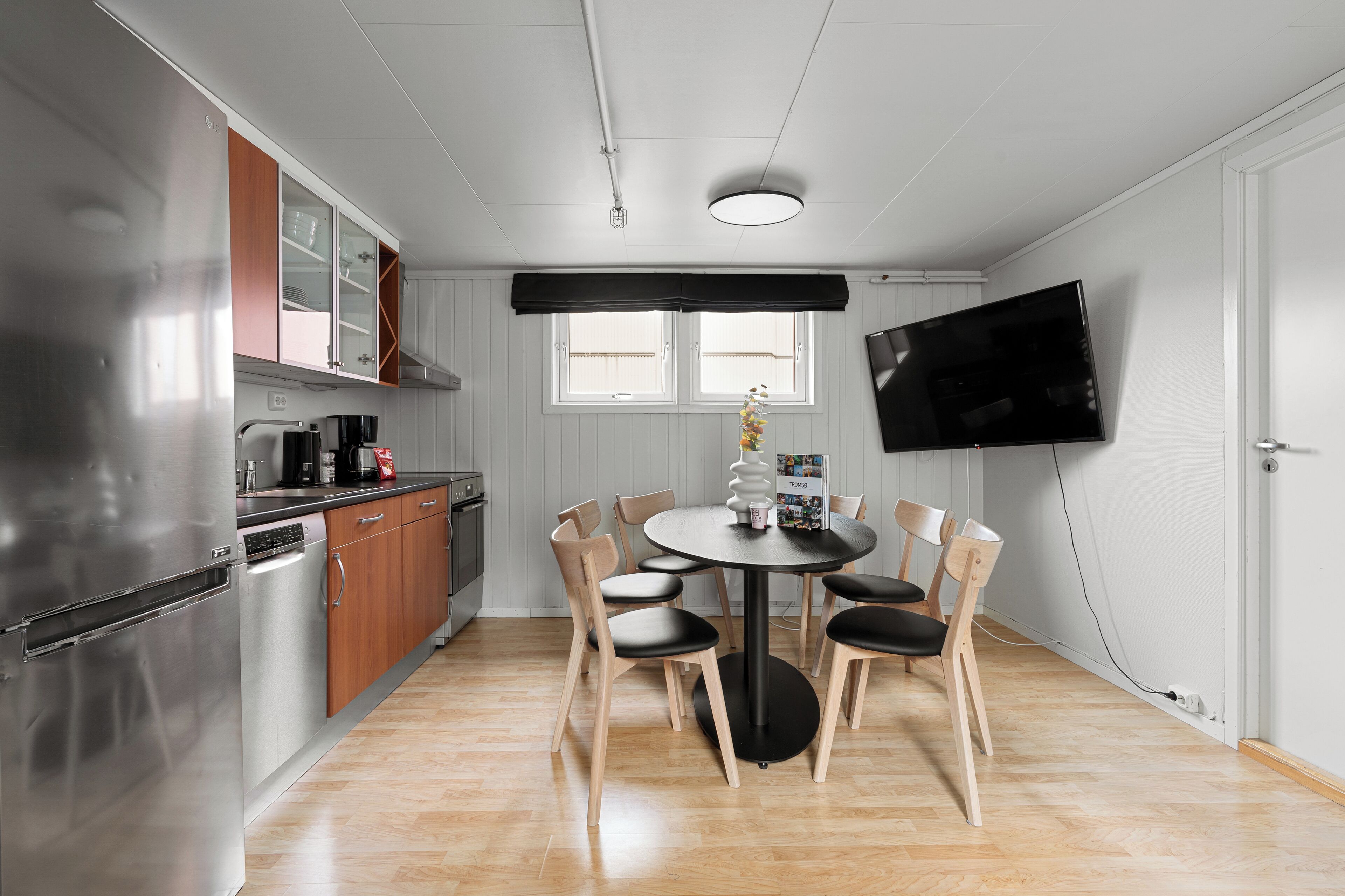 Una cocina moderna con electrodomésticos de acero inoxidable, armarios de madera, una mesa de comedor con sillas y un televisor de pantalla plana montado en la pared.