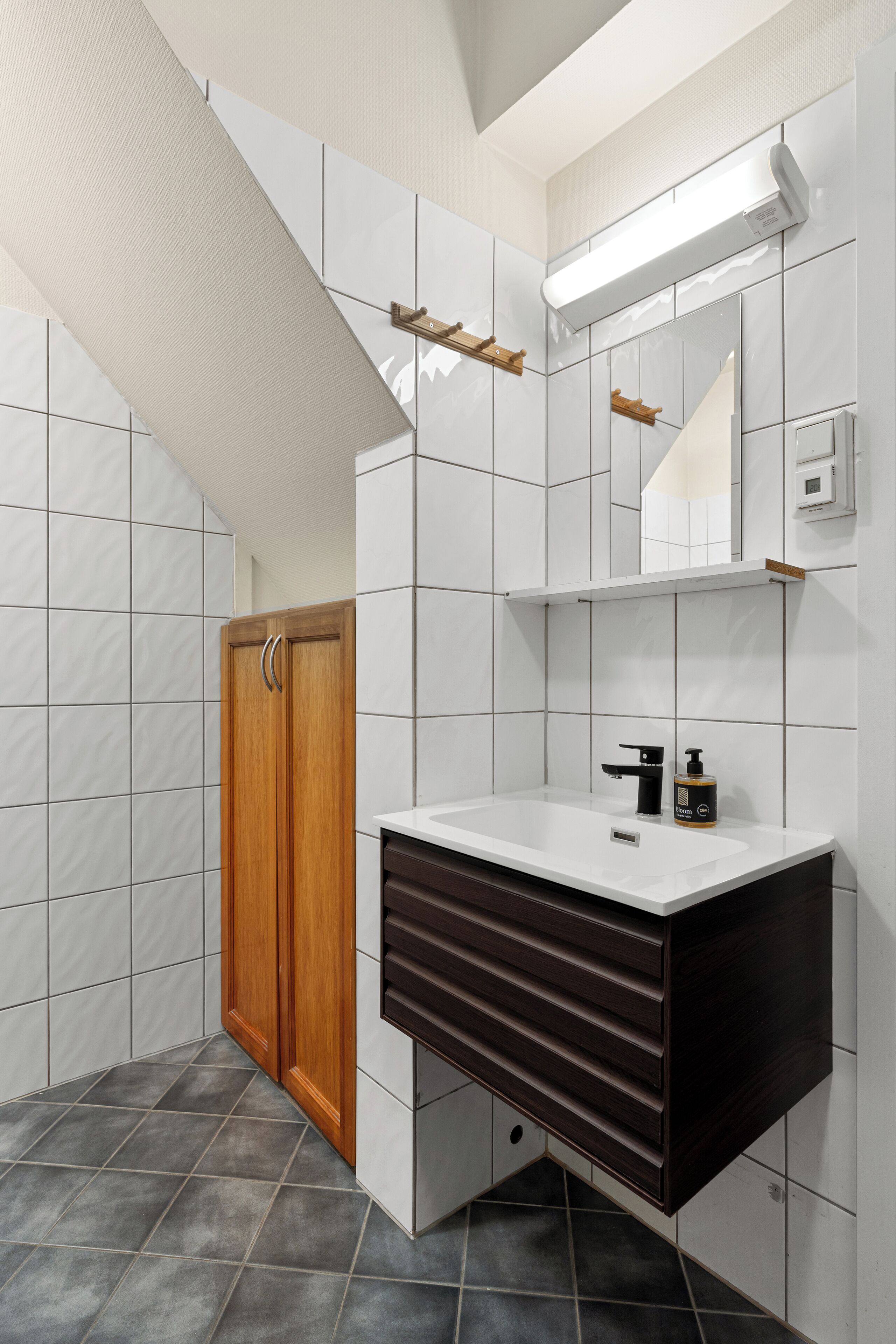 A bathroom with white tiled walls, a wooden door, a dark wooden vanity, a white sink, a mirror, and a towel rack.