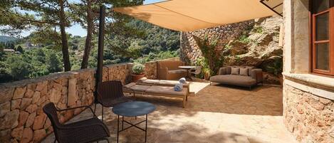 A patio with stone walls, a round table, two chairs, a sofa, and a large umbrella providing shade.