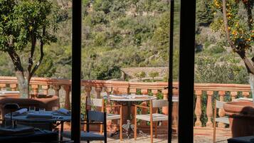 Un área de comedor al aire libre con mesas y sillas, vista a un paisaje montañoso y una barandilla de piedra.