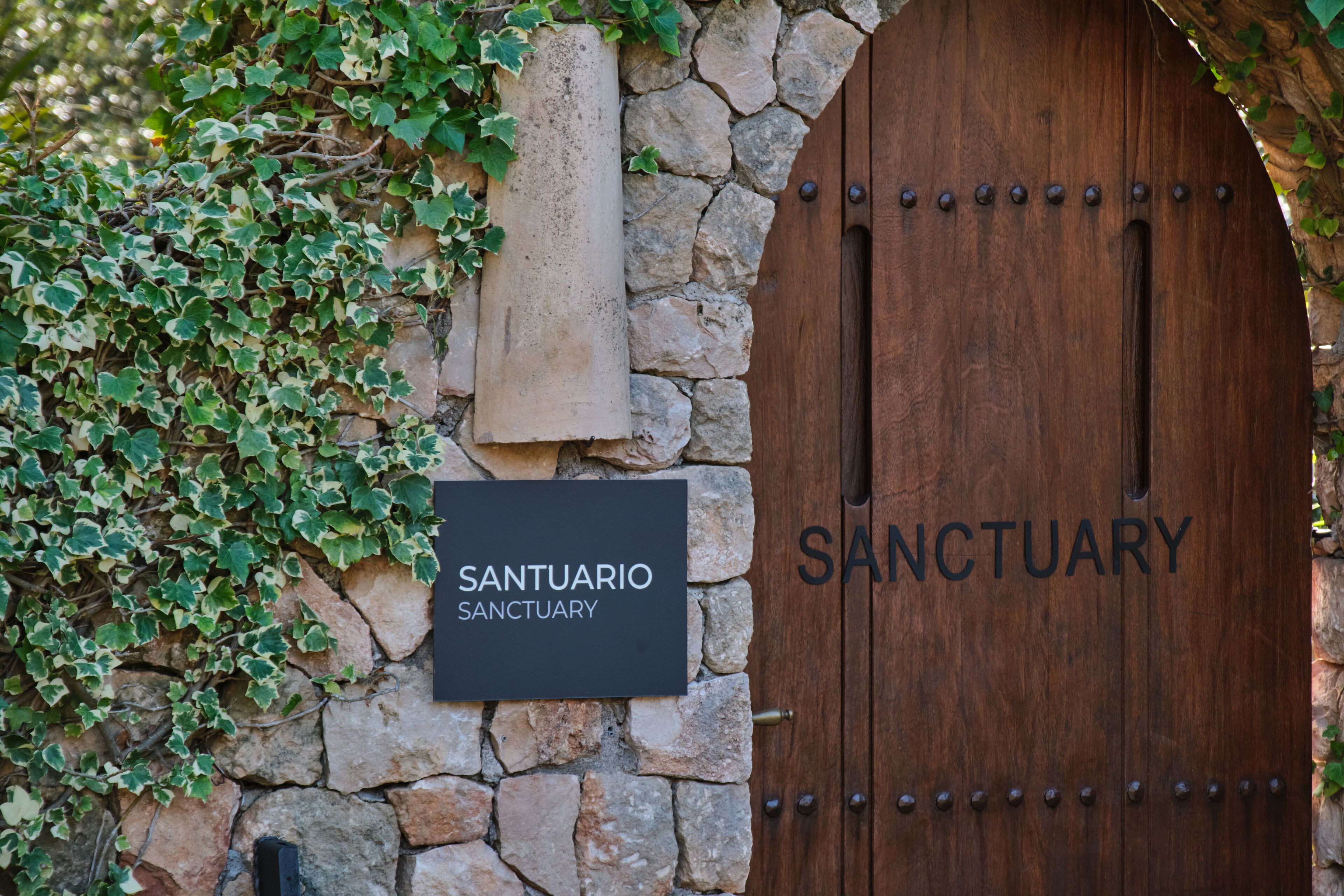 A stone wall with a wooden door labeled "Santuario Sanctuary" and a signboard.