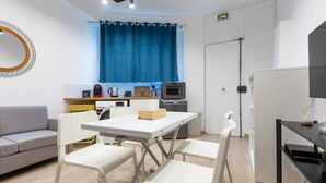 A modern room with a grey sofa, a white table with chairs, a blue curtain, a microwave, a coffee maker, and a small TV. - Home Voltaire (Paris)